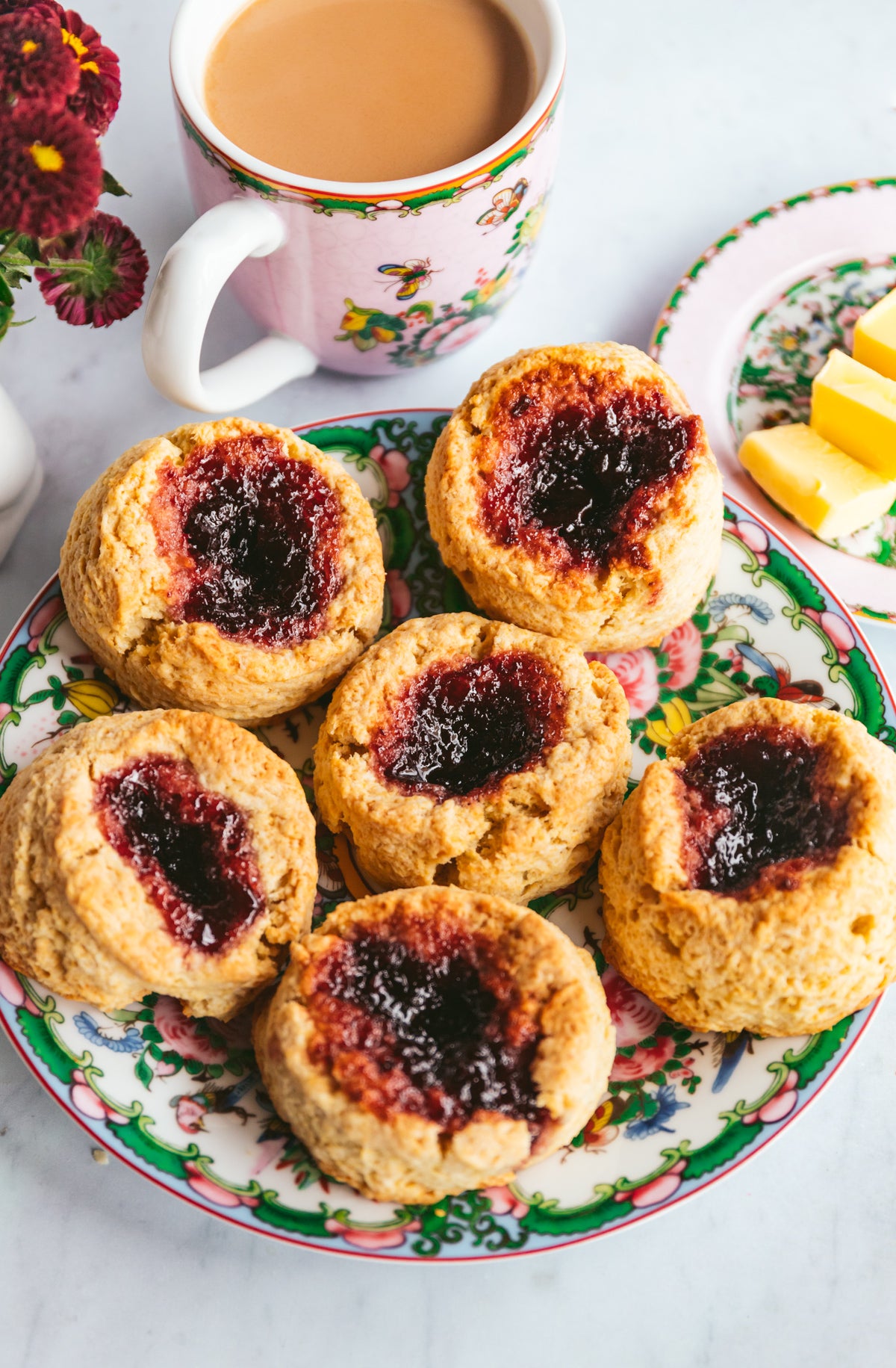 Six golden-brown scones with dark red jam centers arranged on a floral ceramic plate, with a cup of tea, flowers, and butter in a styled breakfast setting.