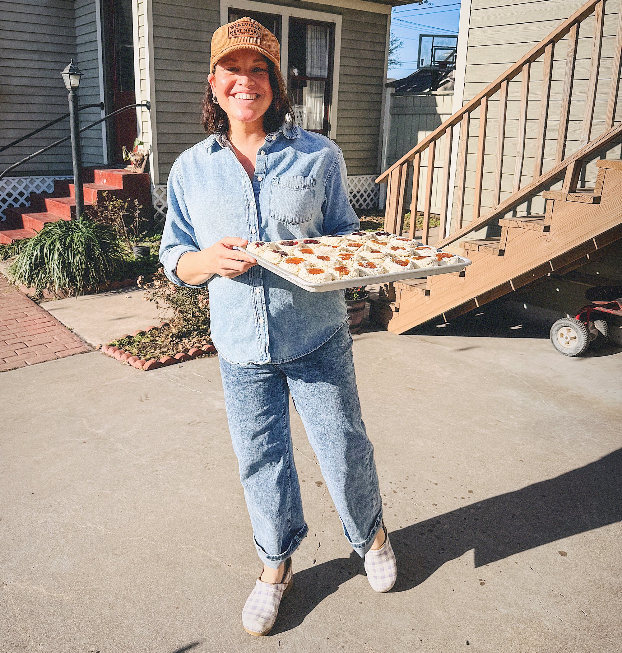 Joy stands outdoors on a concrete driveway in front of a house, holding a metal tray filled with small pastries. She is wearing a light blue denim shirt and jeans, a tan baseball cap, and light-colored slip-on shoes.