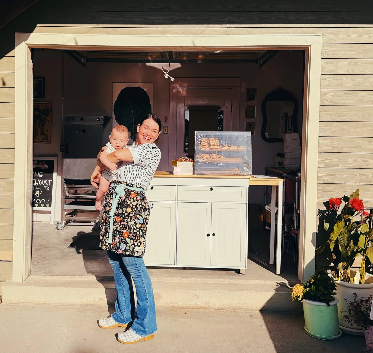 A Joy wearing a floral apron and blue jeans stands in front of an open garage door holding her baby, with a bakery display case and potted flowers visible in the background.